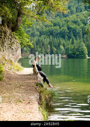 Schöne, süße schwangere Frau sitzt in der Nähe von einem Bergsee in einem Wald am Ufer Stockfoto