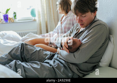 Vater mit Baby im Bett, Frau auf Hintergrund Stockfoto