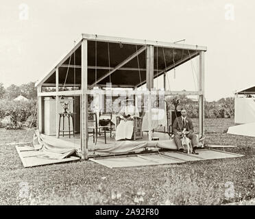 Ein altes Foto von einem TB "frische Luft heilen' Hütte C. 1910. In diesem Sommer - Haus - Stil tented Hütte (Chalet, Hütte oder Kabine) wurde von der Leinwand und Holz auf Holz Rahmen. Es könnte die frische Luft und Sonnenschein in bei gleichzeitigem Schutz der Patient vom Wind und Regen. Vor Penicillin angekommen Tuberkulose (TB) war eine tödliche Krankheit. Eine "Heilung" war frische Luft, Sonne und Ruhe. Es ist zusammenklappbar Modus - seine Seiten abgesenkt extra frischen Wind in die Hütte zu ermöglichen. Im Inneren befindet sich ein Bett, Stühle und Schränke. Die Frau trägt die Uniform eine Krankenschwester und beide Sie und der Mann haben einen Hund und Welpen gebracht. Stockfoto