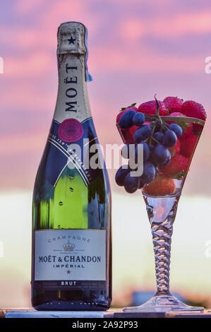 Eine Flasche Champagner und ein Glas Champagner mit Trauben und Obst gegen Sonnenuntergang Himmel Stockfoto