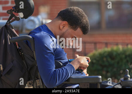 Afroamerikanischer Mann mit Zerebralparese mit Handy Auf seinem Power-Rollstuhl draußen Stockfoto