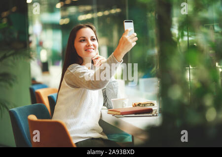 Portrait der junge lächelnde Schüler Mädchen machen Video Anruf vom Smartphone zu Ihrem besten Freund, in Pause, gute Nachrichten, sitzt im Café, funktioniert Stockfoto