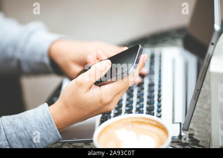 Nahaufnahme der Laptop auf dem Tisch. Mädchen in beige Pullover bei Tisch holding Smartphone sitzen. Frau entscheidet, Produkte im Online Store verwendet Computer. Eine Frau prüft die E-Mails beim Sitzen in Cafés. Stockfoto