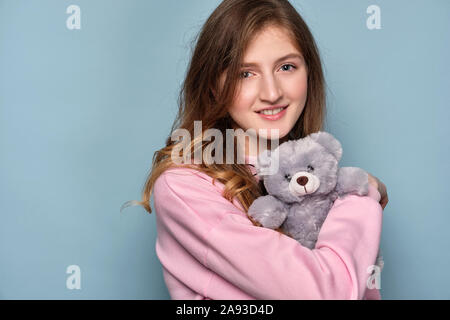 Blue-Eyed Girl cuddles mit einem Spielzeug Grauer Bär, stehend auf einem blauen Hintergrund. Stockfoto