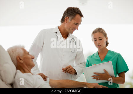 Älterer Mann liegt im Krankenhausbett, während Arzt und Krankenschwester Sprechen Sie an seinem Bett Stockfoto