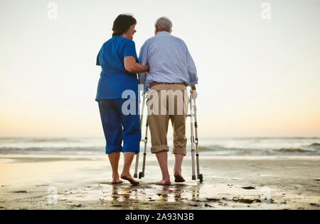 Älterer Mann, der mit einer Gehhilfe neben einem läuft Krankenschwester am Strand Stockfoto