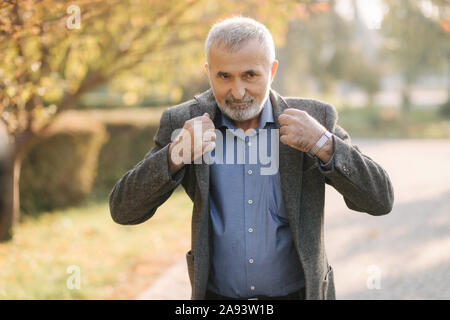 Gut aussehender älterer Mann setzen auf eine graue Jacke. Alte grauhaarige bärtiger Mann Spaziergang im Herbst Park. Gelber Hintergrund Stockfoto