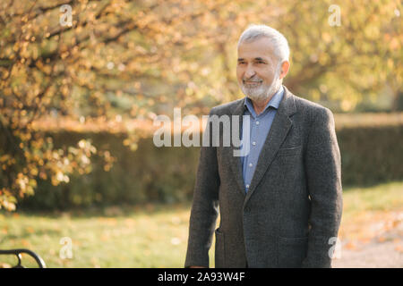 Gut aussehender älterer Mann setzen auf eine graue Jacke. Alte grauhaarige bärtiger Mann Spaziergang im Herbst Park. Gelber Hintergrund Stockfoto