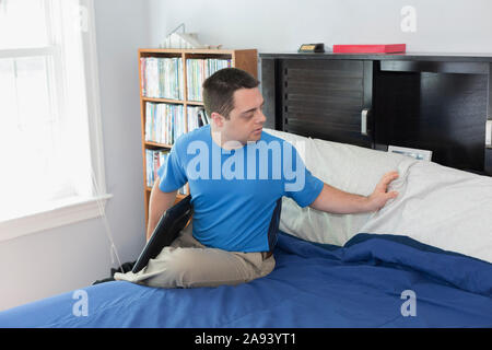 Mann mit Down-Syndrom sitzt auf dem Bett und hält einen Laptop Stockfoto