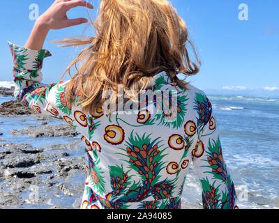 Junge rötlich-blonde Frau von hinten gesehen, mit geblümten Kleid, Wind im Haar, mit Blick auf das Meer vom Ufer Stockfoto