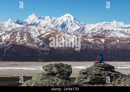 Ein Wanderer blickt auf den McGinnis Peak, Mount Moffit und Mount Hayes in der Eastern Alaska Range, während er sich auf einem Felsen über dem Delta ausruht... Stockfoto