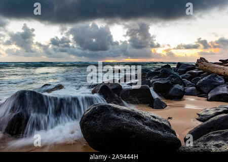 Surfen Waschen über Felsen am Strand, wie die Sonne über dem Meer mit dunklen Wolken über; Kapaa, Kauai, Hawaii, Vereinigte Staaten von Amerika Stockfoto