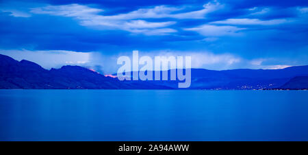 Ein Waldbrand brennt in den Bergen in der Ferne mit dem Okanagan-See im Vordergrund unter einem launischen, stürmischen Himmel; British Columbia, Kanada Stockfoto