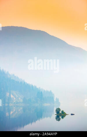 Dichter Nebel über einem See und einem Berg mit einem leuchtend orangefarbenen Himmel bei Sonnenaufgang; British Columbia, Kanada Stockfoto