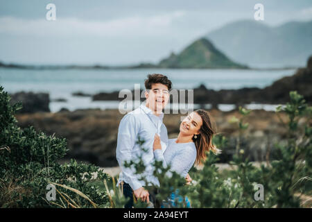 Porträt eines jungen Paares mit der zerklüfteten Küste von North Island im Hintergrund; Wellington, North Island, Neuseeland Stockfoto