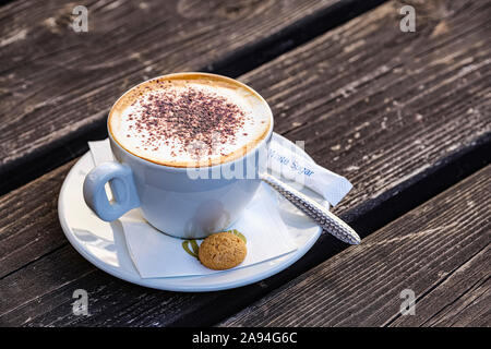 Cappuccino von einem italienischen Café auf Holztisch, serviert mit einem kleinen Keks; Hexham, Northumberland, England Stockfoto