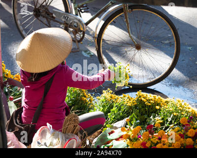 Vietnamesische Frau mit einer konischen Strohhut oder Chrysanthemen la und ein rosa Kapuzenpulli Verkauf von Gold und Rot an einer im Markt in Hoi An Stockfoto