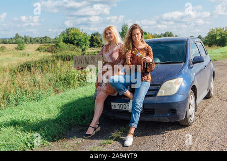 Zwei hübsche Mädchen per Anhalter und mit einem Zeichen überall sitzen auf der Motorhaube eines Autos am Straßenrand stimmen. Stockfoto
