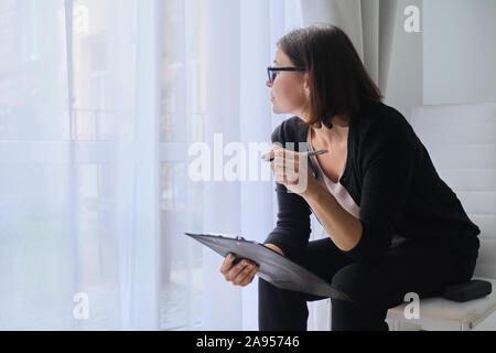 Serious Business reife Frau mit Zwischenablage sitzen in der Nähe der Fenster Stockfoto