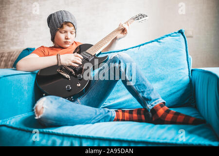 Stilvolle Rock und Roll Kid mit einem schwarzen E-Gitarre sitzt auf einem Stuhl und spielt Musik Stockfoto