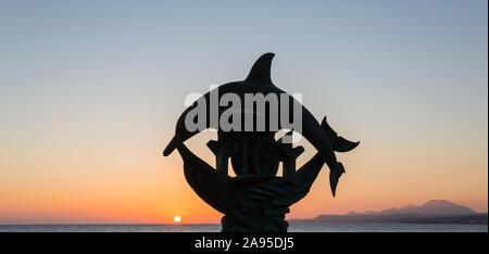 Rethymno, Kreta, Griechenland. Silhouette der ikonischen Delphin Statue bei Sonnenaufgang, Mount Psiloritis im Hintergrund sichtbar. Stockfoto