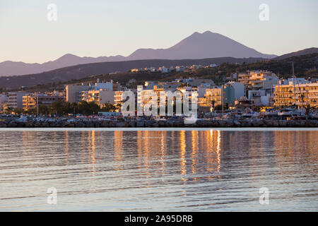 Rethymno, Kreta, Griechenland. Gebäude am Meer spiegeln sich in stillem Wasser, Sonnenaufgang, Mount Psiloritis sichtbar darüber hinaus. Stockfoto