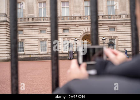 Ein männlicher Tourist vor den Toren des Buckingham Palace fotografiert mit seinem Handy von Queen's Guard, das im Dienst in den Sentry-Boxen steht, die den königlichen Monarchen bewachen. Stockfoto