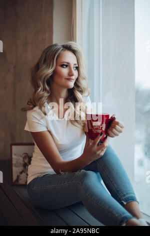 Schöne Blondine sitzen auf Fensterbank mit Becher. Stockfoto