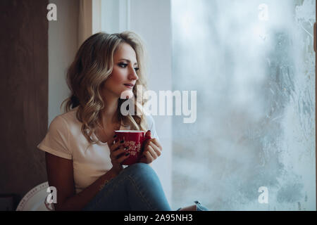 Schöne Blondine sitzen auf Fensterbank mit Becher. Stockfoto