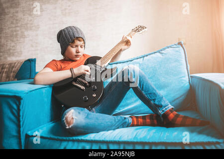 Informelle rothaarige Teenager in Jeans, hat und Red t-shirt spielt Rock auf einem schwarzen E-Gitarre auf dem Sofa zu Hause sitzen Stockfoto