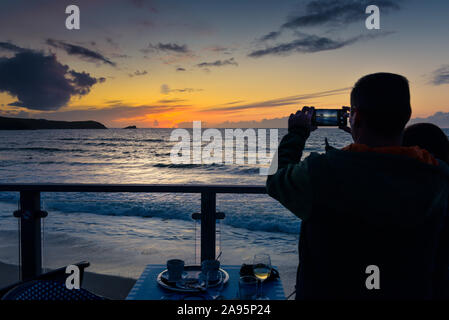 Einen Urlauber mit einem Smartphone eine schöne intensive Sonnenuntergang auf den Fistral Bay in Newquay in Cornwall zu fotografieren. Stockfoto