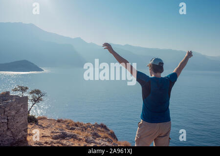 Junge Mann steht auf der Oberseite mit ausgestreckten Armen, gegen das Meer und den Horizont. Ansicht von hinten. Stockfoto