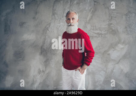 Foto von attraktiven alter Kerl, Hände in den Taschen selbstbewusst tragen rote Strickpullover weiße Hose coole Hipster santa Kleidung grau isoliert Stockfoto