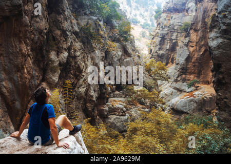 Ein Reisender in Einheit mit der Natur, ein junger Mann sitzt auf einem Felsen in einer felsigen Schlucht und blickt voraus Stockfoto