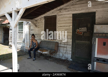 Asiatische Frau in Sonnenbrille sitzt auf der Bank in verlassenen Tankstelle in Virginia Stockfoto