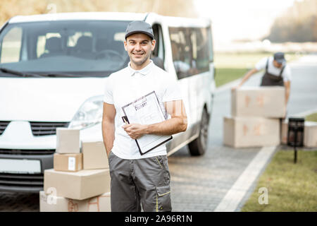 Porträt der Mann in Uniform stehend mit Dokumenten, die in der Nähe der Cargo van Fahrzeug, Kollege entlädt Pakete auf dem Hintergrund Stockfoto