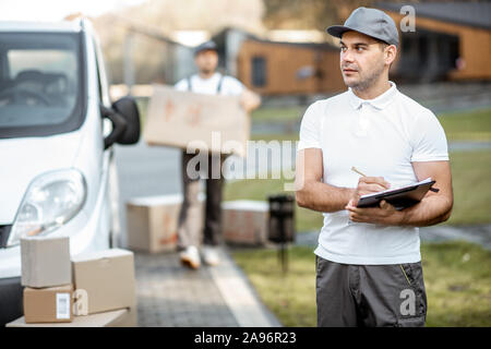 Porträt der Mann in Uniform stehend mit Dokumenten, die in der Nähe der Cargo van Fahrzeug, Kollege entlädt Pakete auf dem Hintergrund Stockfoto