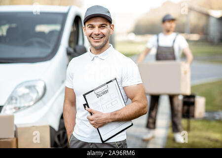 Porträt der Mann in Uniform stehend mit Dokumenten, die in der Nähe der Cargo van Fahrzeug, Kollege entlädt Pakete auf dem Hintergrund Stockfoto