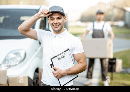 Porträt der Mann in Uniform stehend mit Dokumenten, die in der Nähe der Cargo van Fahrzeug, Kollege entlädt Pakete auf dem Hintergrund Stockfoto