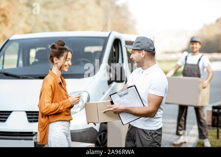 Kurier mit Checkliste Ware liefern, um eine junge Frau, die durch die Ladung van Fahrzeug, Mover mit Karton Pakete auf dem Hintergrund Stockfoto