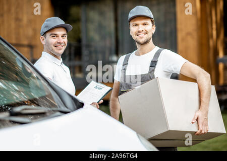 Porträt einer zwei freundliche Lieferung Männer in Uniform stehen zusammen mit Checkliste und Kartons in der Nähe der Cargo van Fahrzeug im Freien Stockfoto