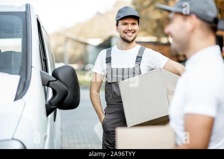 Zwei fröhliche männlichen Kuriere in Uniform Lieferung von Cargo van Fahrzeug, entladen Karton Pakete von einem Auto im Freien Stockfoto