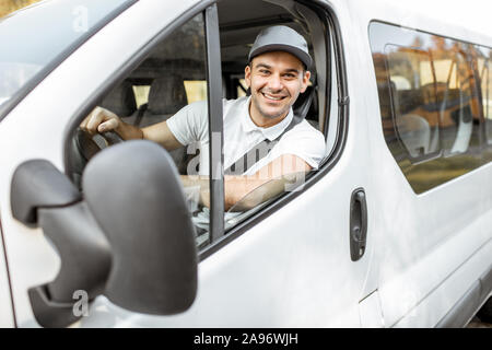 Porträt einer fröhlichen Anlieferung Treiber in Uniform Blick aus dem Fenster des weißen Lieferwagen vahicle, Lieferung mit dem Auto Stockfoto