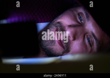 Bärtiger junger Mann im Bett liegend unter seiner Decke. Er kann nicht schlafen und ist etwas aufpassen auf sein Mobiltelefon. Stockfoto