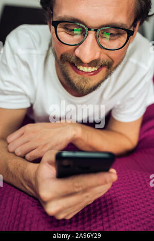 Schön lächelnden jungen Mann mit Brille liegend auf Bett, während sein Handy zu halten. Stockfoto