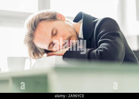 Hübscher junger Mann schlafen auf Stapel Bücher am Schreibtisch. Kaffeetasse auf dem Schreibtisch neben ihm sichtbar. Stockfoto