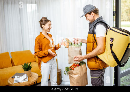 Courier in Uniform mit thermischen Tasche liefert frische Lebensmittel in Tüten zu einem jungen Client Home. Glückliche Frau warten auf der Bestellung Stockfoto