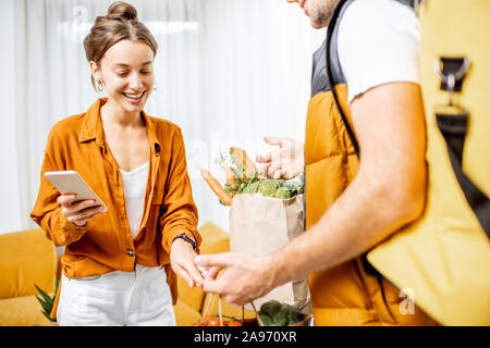 Courier in Uniform mit thermischen Tasche liefert frische Lebensmittel in Tüten zu einem Client Home. Frau mit Smartphone Kontrolle ihrer Bestellung Stockfoto