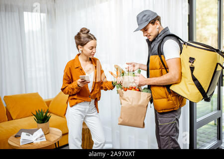 Courier in Uniform mit thermischen Tasche liefert frische Lebensmittel in Tüten zu einem Client Home. Frau mit Smartphone Kontrolle ihrer Bestellung Stockfoto