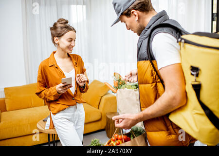 Courier in Uniform mit thermischen Tasche liefert frische Lebensmittel in Tüten zu einem Client Home. Frau mit Smartphone Kontrolle ihrer Bestellung Stockfoto